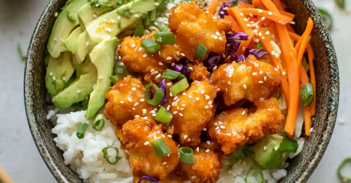 Bang Bang Chicken Bowl with crispy chicken, rice, shredded cabbage, carrots, avocado, and creamy bang bang sauce, garnished with green onions and sesame seeds