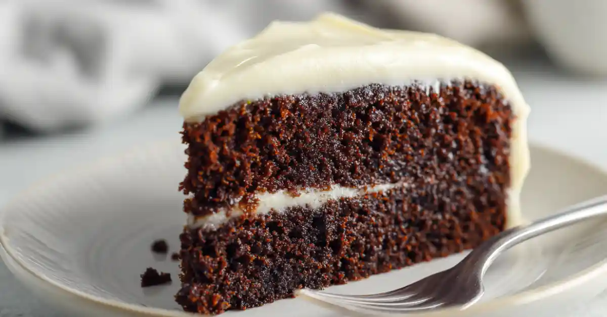 Moist chocolate zucchini cake slice with cream cheese frosting on a white plate in a minimal kitchen setting.