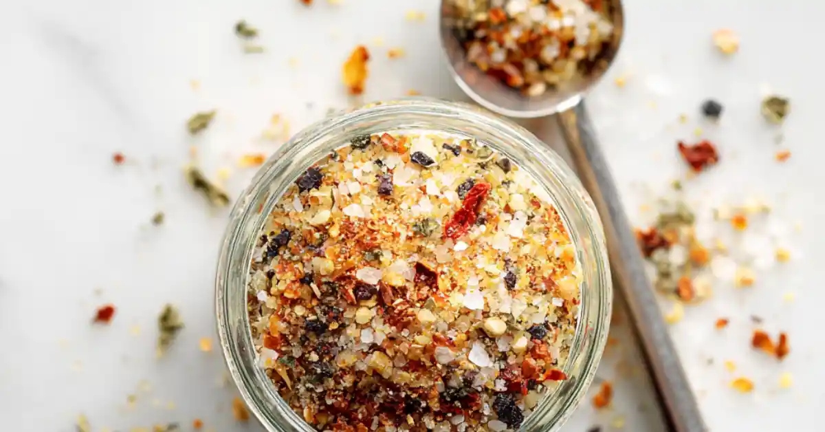 Homemade Montreal steak seasoning in a glass jar on a white marble surface with a measuring spoon beside it.